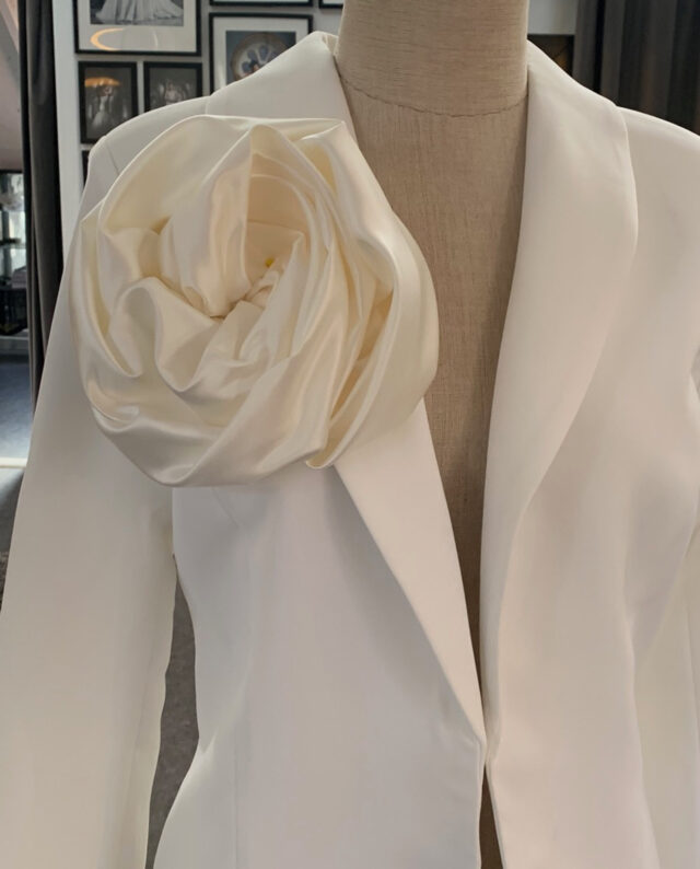 Satin Rosette – Ivory A soft, sculptural satin rosette in timeless ivory. Each piece is handmade and finished with a brooch pin on the back, making it easy to attach to dresses, blazers, or belts for a refined and eye-catching touch.